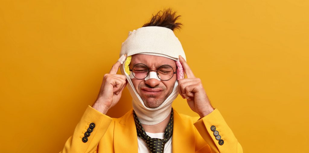 Displeased man suffers from unbearable migraine after injury, dressed in formal clothes, has bruises and broken nose, recovers after difficult surgery operation, isolated on yellow background