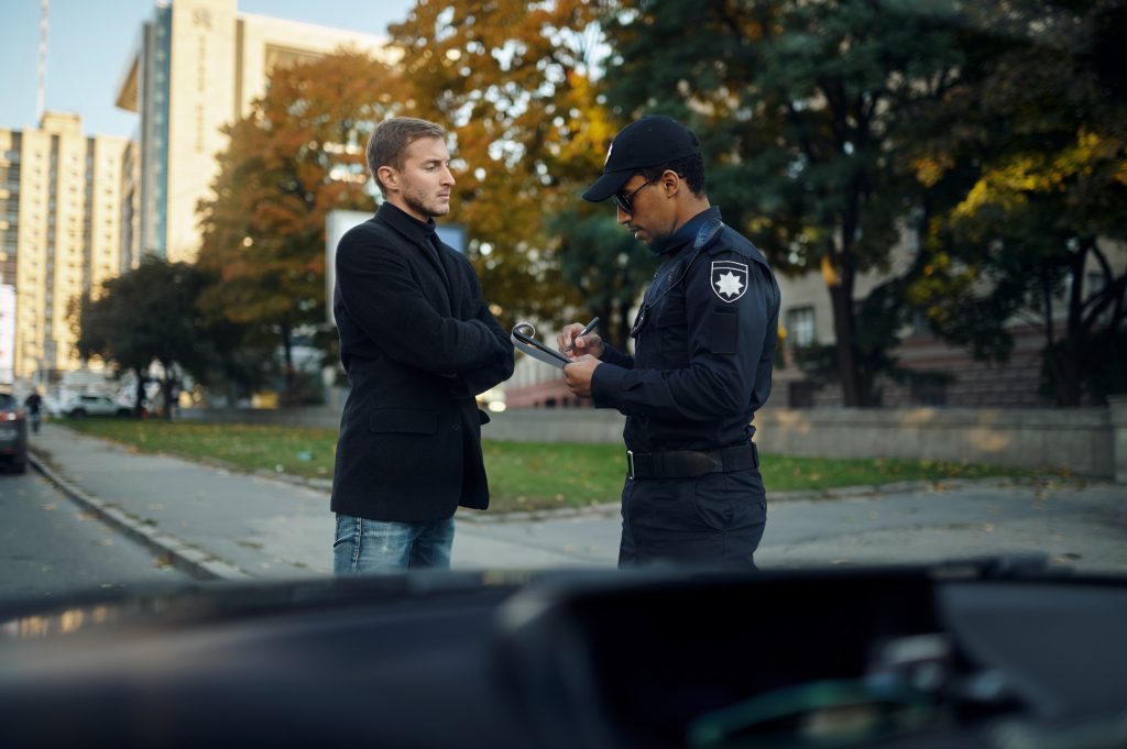 A man in a dark coat speaking with a police officer who is writing on a clipboard, with the blurred outline of a car in the foreground and autumn trees in the background, depicting a typical post-accident scene on a city street.